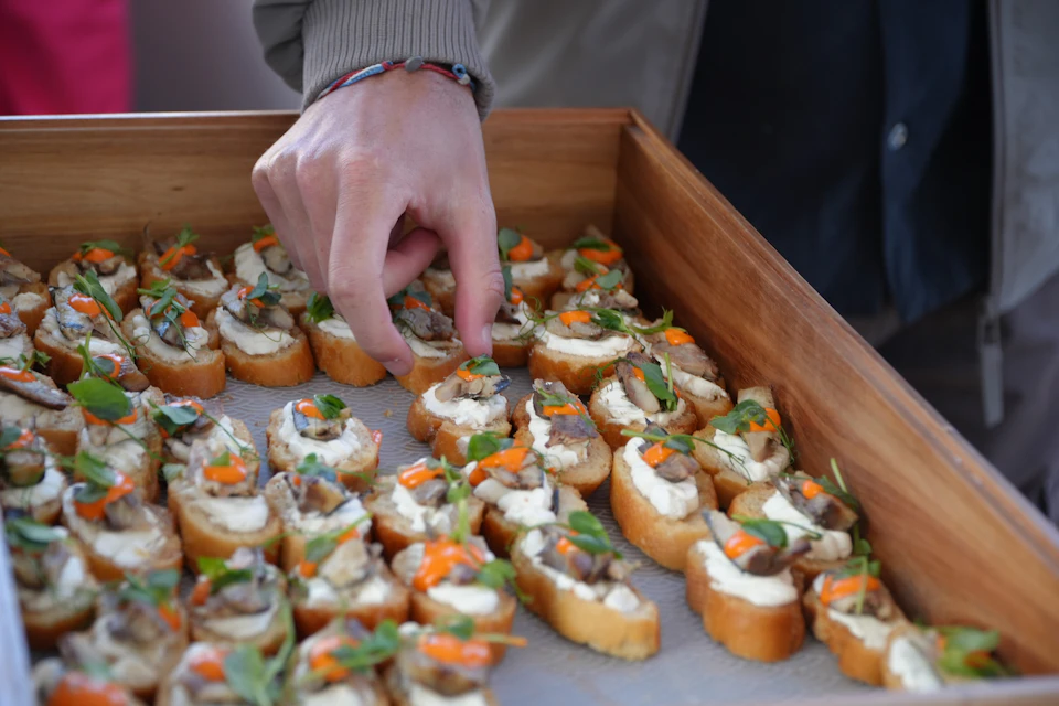 A hand reaching for a small appetizer piece from a wooden tray filled with decorated bread slices with toppings.
