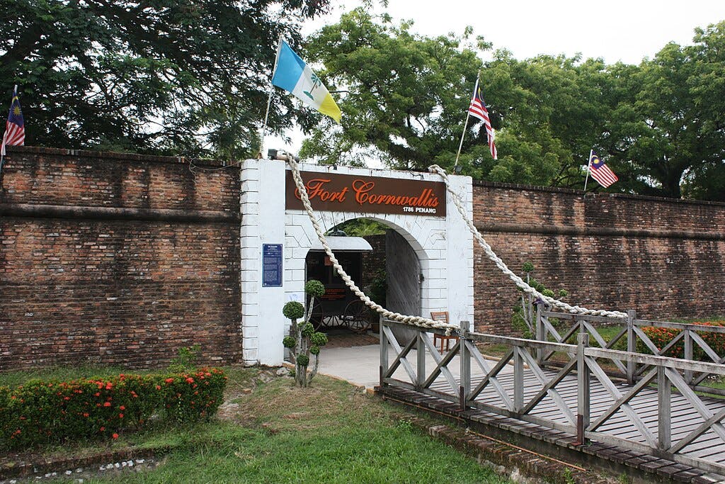 Entrance of Fort Cornwallis featuring a brick wall, arched gate, flags, a wooden drawbridge, and surrounding greenery.