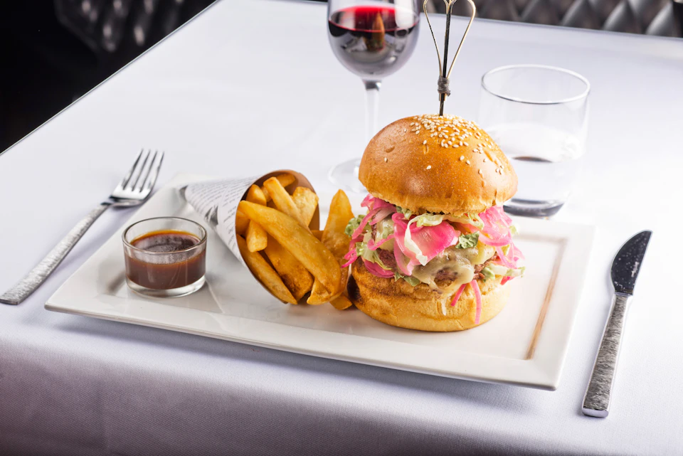 Gourmet burger with pickled onions, fries in a paper cone, and a glass of red wine on a white tablecloth.