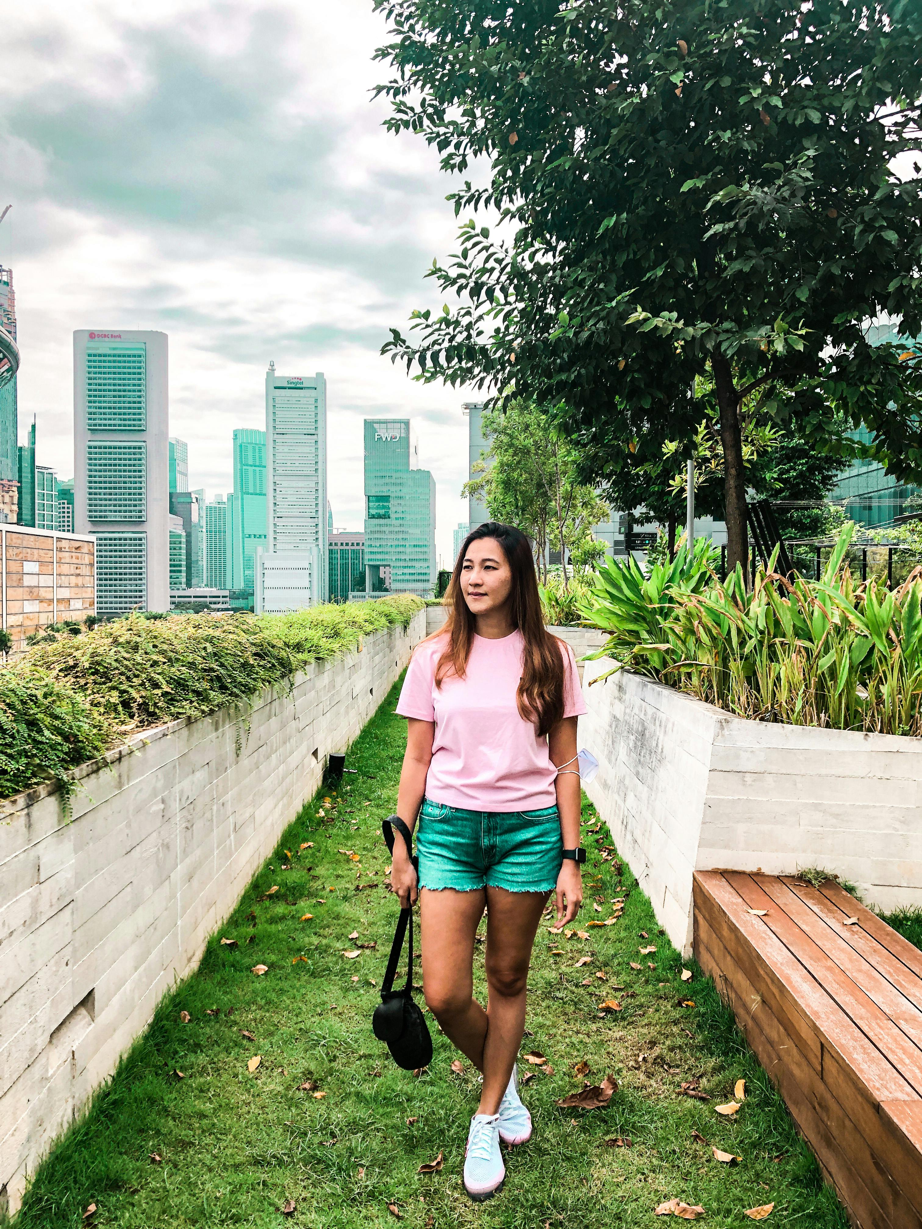 Una donna in maglietta rosa e pantaloncini verdi si trova in un lussureggiante giardino pensile con i grattacieli della città sullo sfondo.