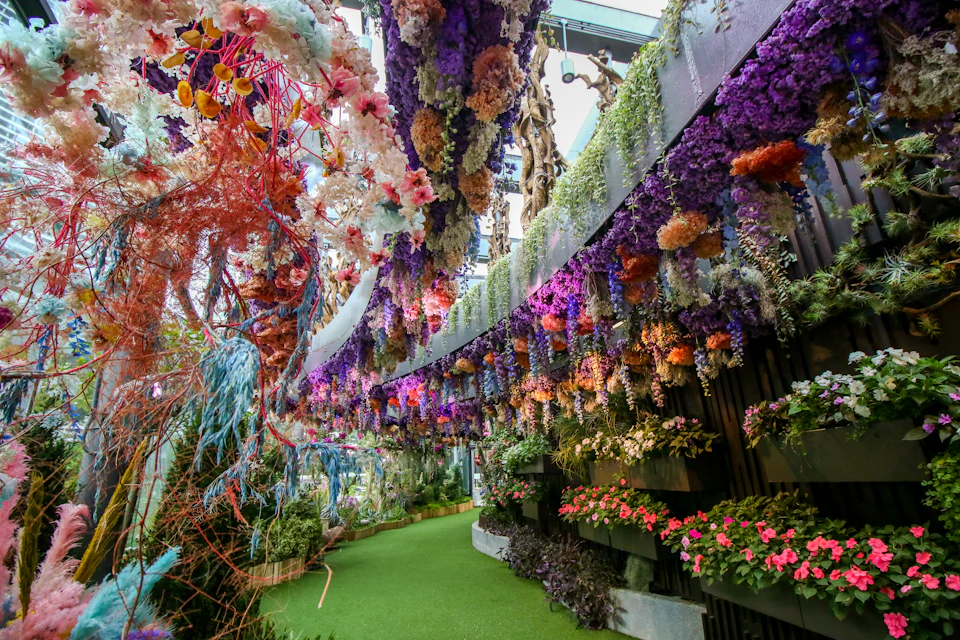A lush, colorful indoor garden with hanging floral arrangements, green artificial turf, and potted plants along the sides.