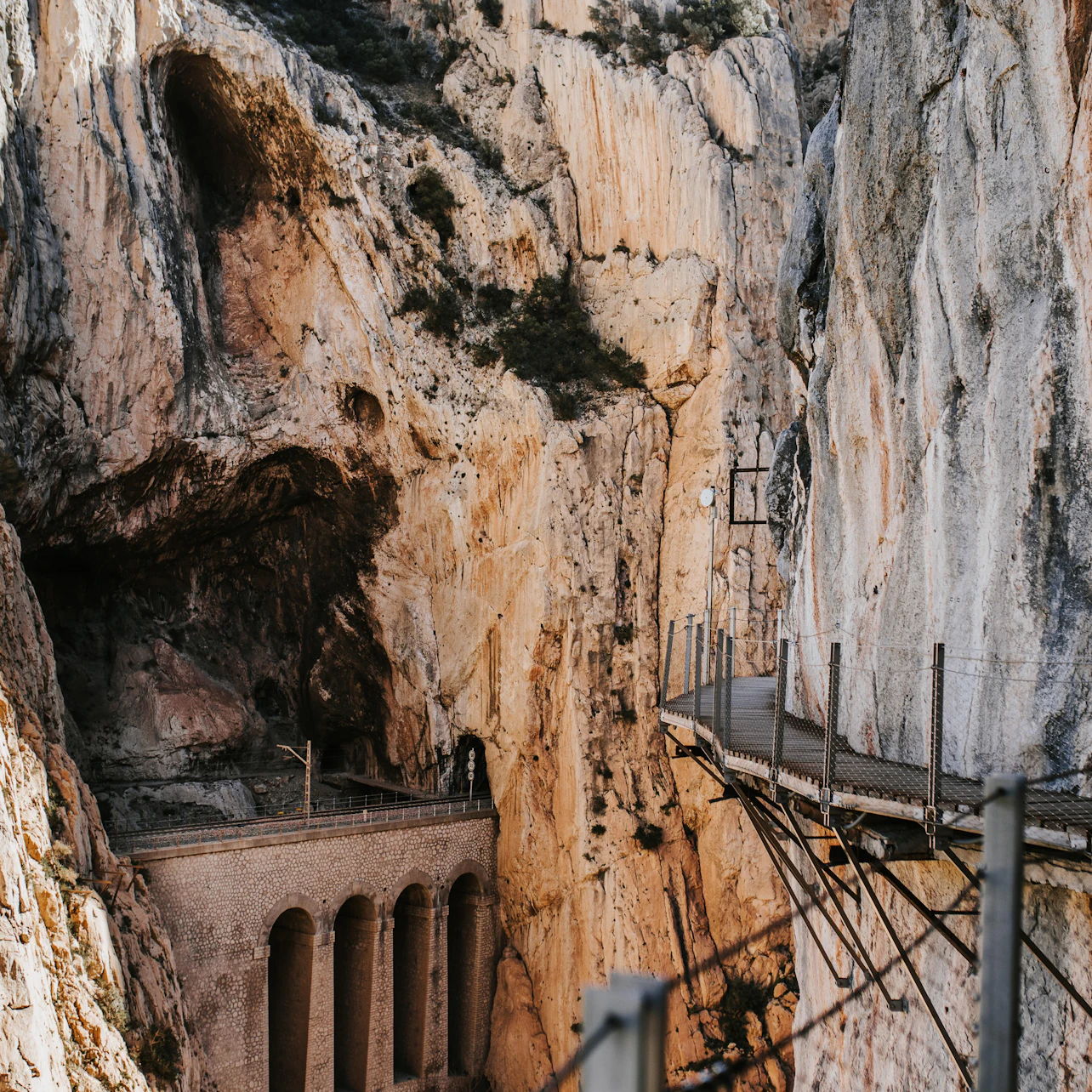 A narrow walking path with railings runs along a steep rocky gorge, with an arched bridge visible below.