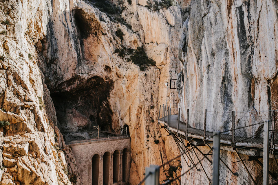 A narrow walking path with railings runs along a steep rocky gorge, with an arched bridge visible below.