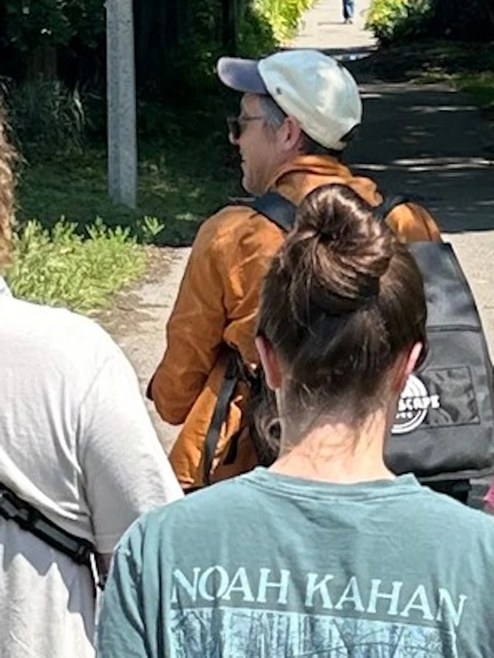 A group of people walking on a paved path outdoors, surrounded by greenery and partial fences under a sunny sky.