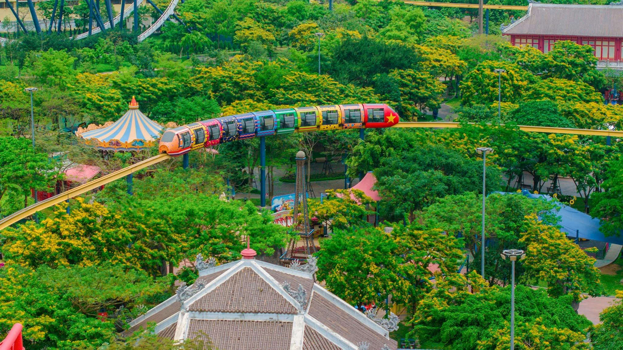 A colorful train on an elevated track in a park surrounded by lush greenery and trees.