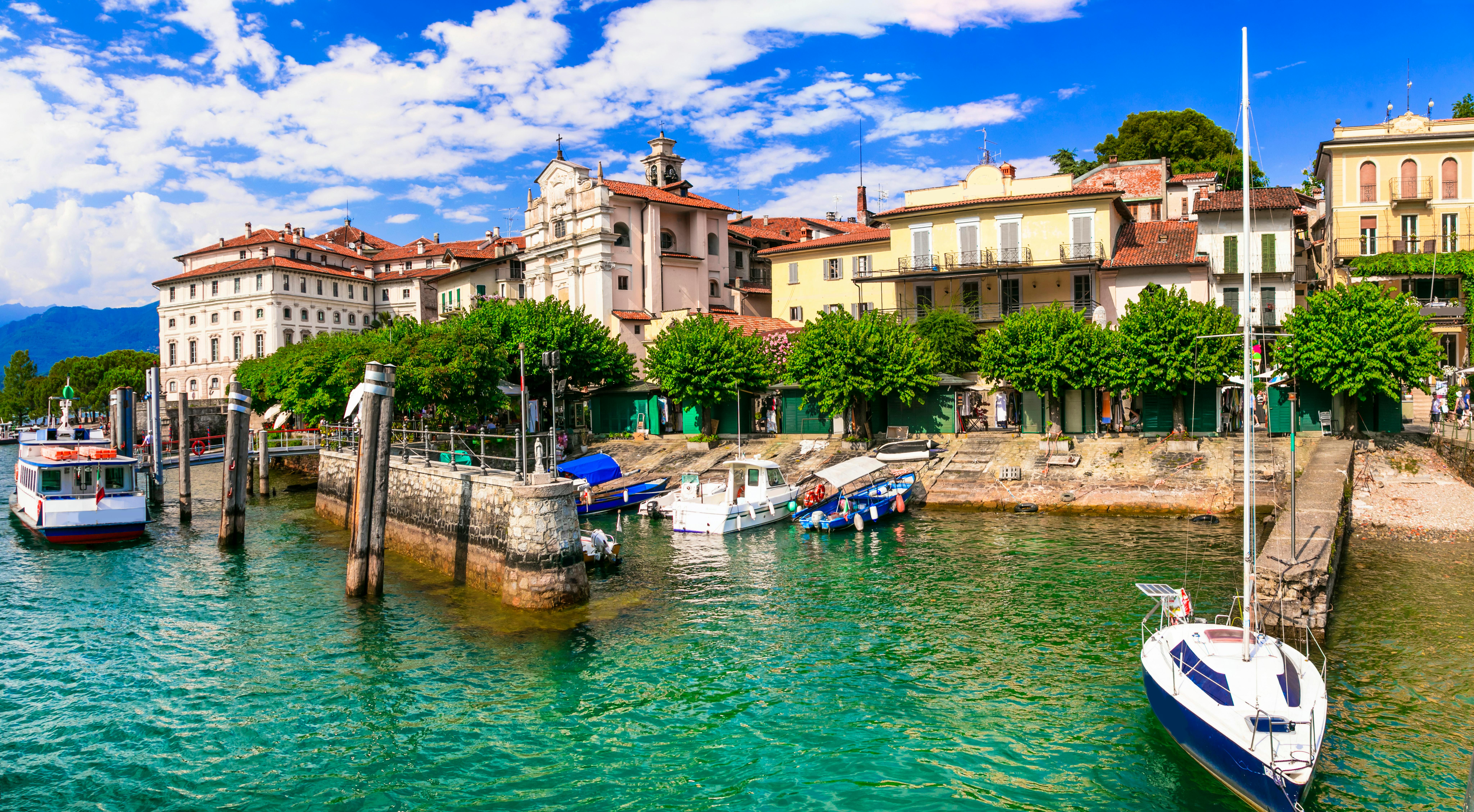 Coastal town with boats docked, historic buildings, trees, and a clear blue sky reflected in green waters.