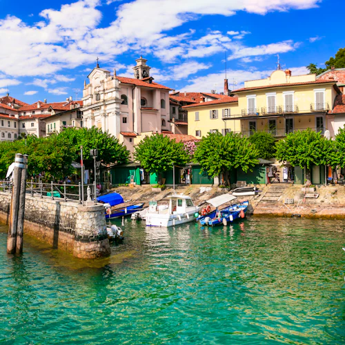 Kuststad med dockade båtar, historiska byggnader, träd och en klarblå himmel som speglas i grönt vatten.