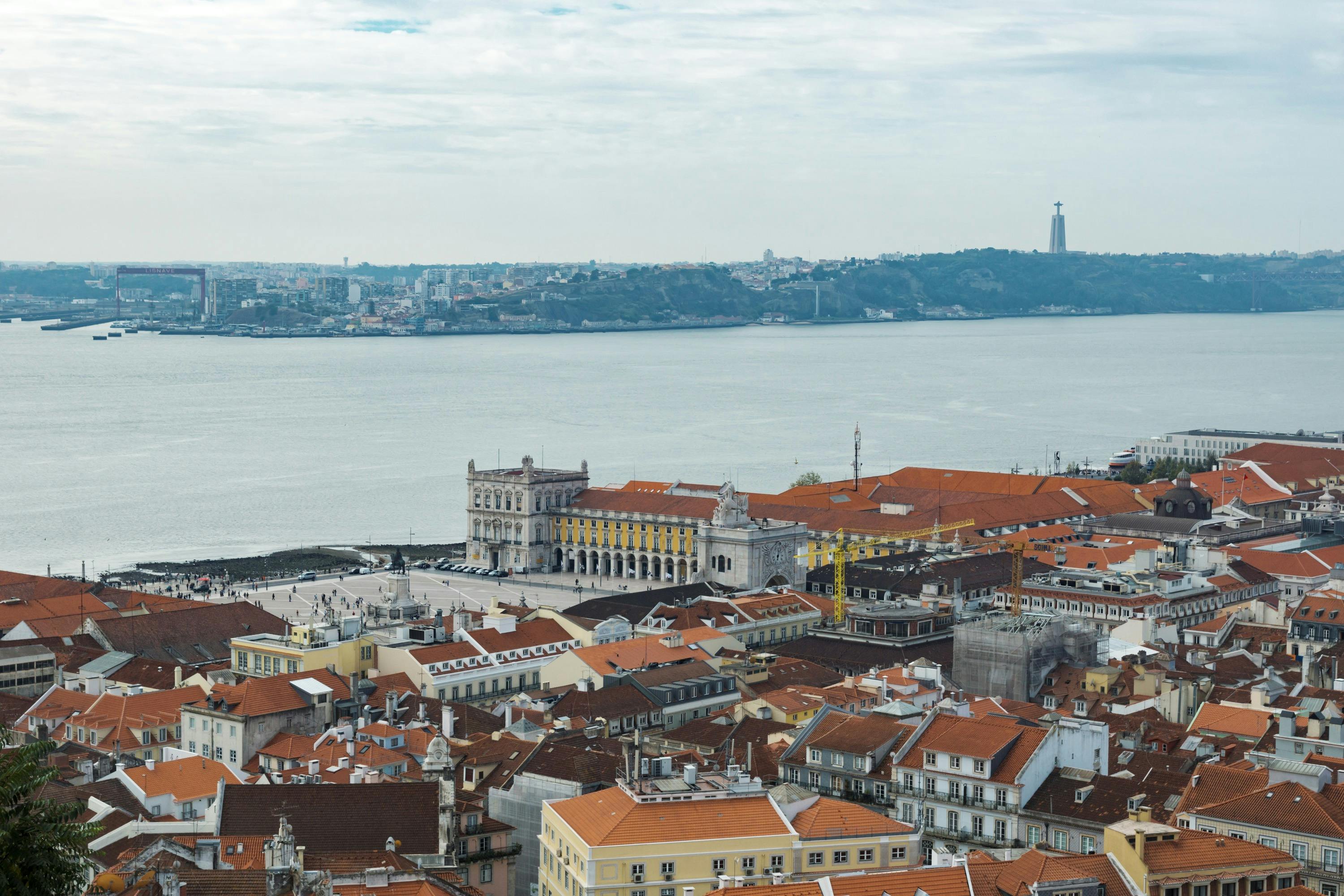 A view of a coastal city with red-roofed buildings, a river, and a statue in the background under a cloudy sky.