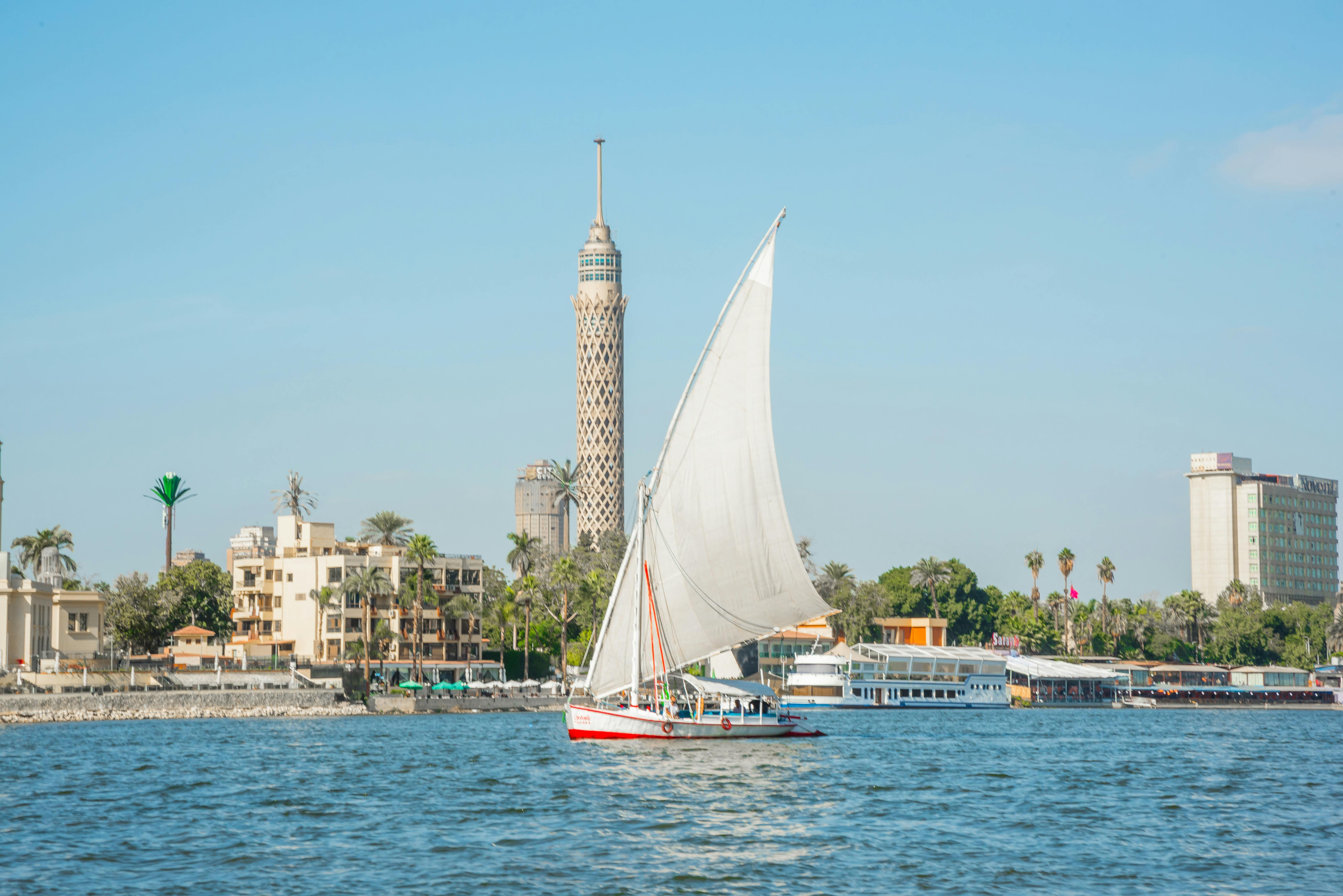 A sailboat with a large white sail on a river, with buildings and a tall tower in the background under a clear blue sky.