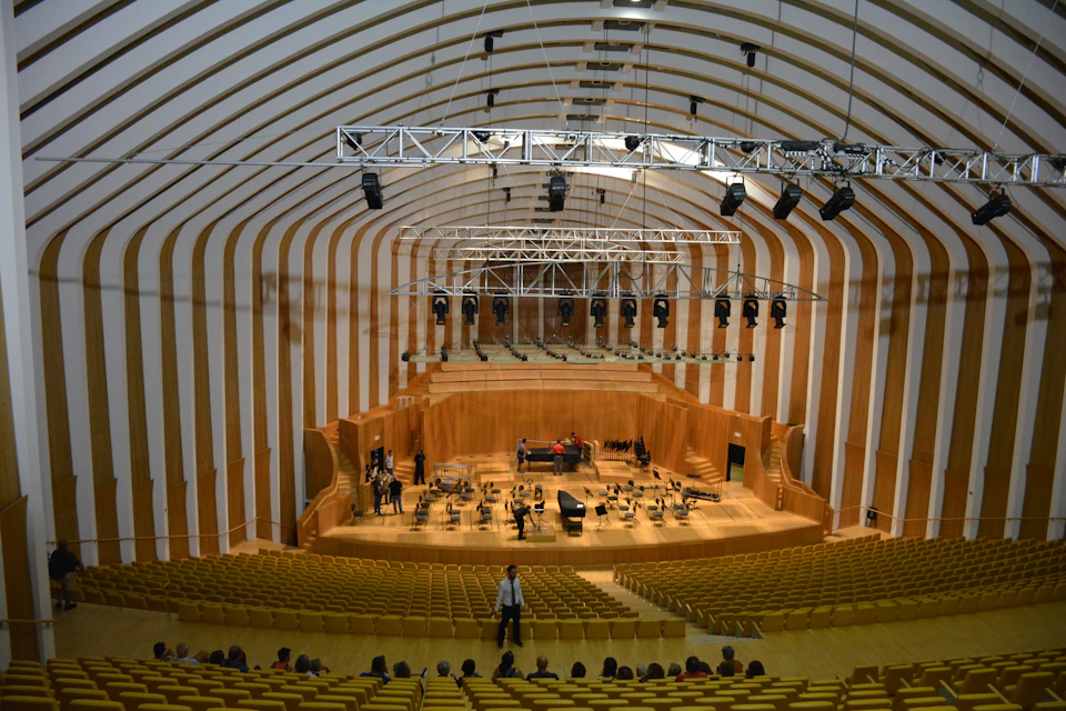 A large, modern auditorium with tiered seating, a stage with musical instruments, and lighting rigs overhead. People are setting up the stage.