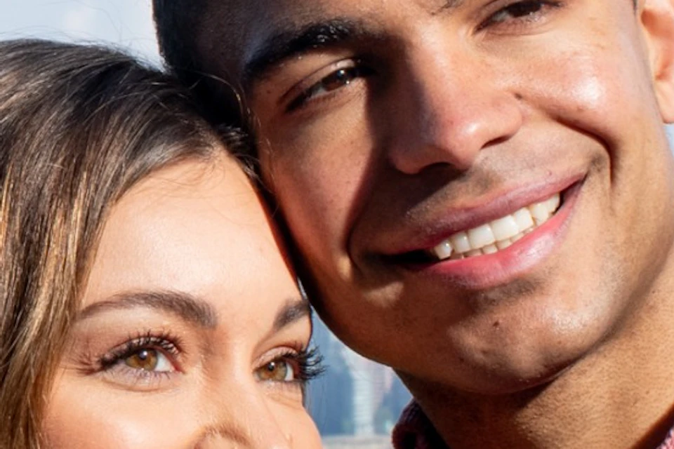 A couple smiles while taking a selfie with a city skyline and green park in the background.