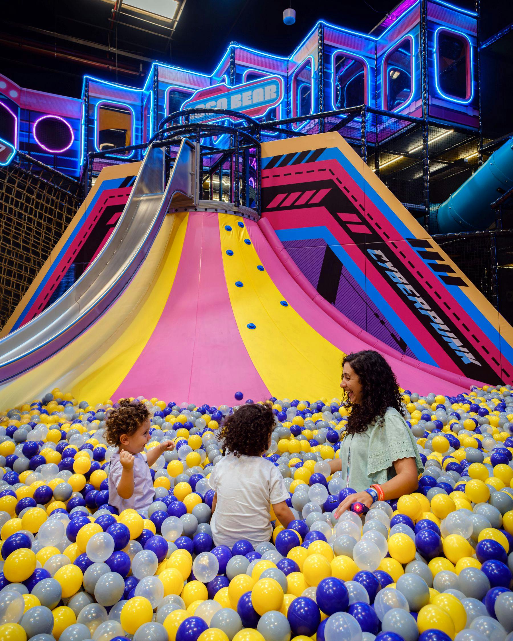 Τρία άτομα, συμπεριλαμβανομένης μιας γυναίκας και δύο παιδιών, παίζουν σε έναν πολύχρωμο λάκκο με μπάλες μπροστά από μια τσουλήθρα σε έναν εσωτερικό χώρο παιχνιδιού.