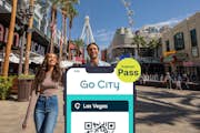 Las Vegas Explorer Pass by Go City es mostra en un telèfon intel·ligent amb una parella de turistes al Strip de Las Vegas al fons