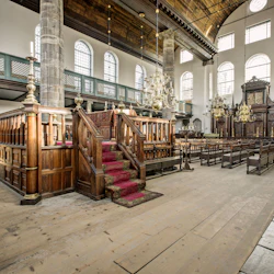 En utsmyckad, tom synagoginteriör med stora välvda fönster, träbänkar, ljuskronor och en bimah med röda mattor.