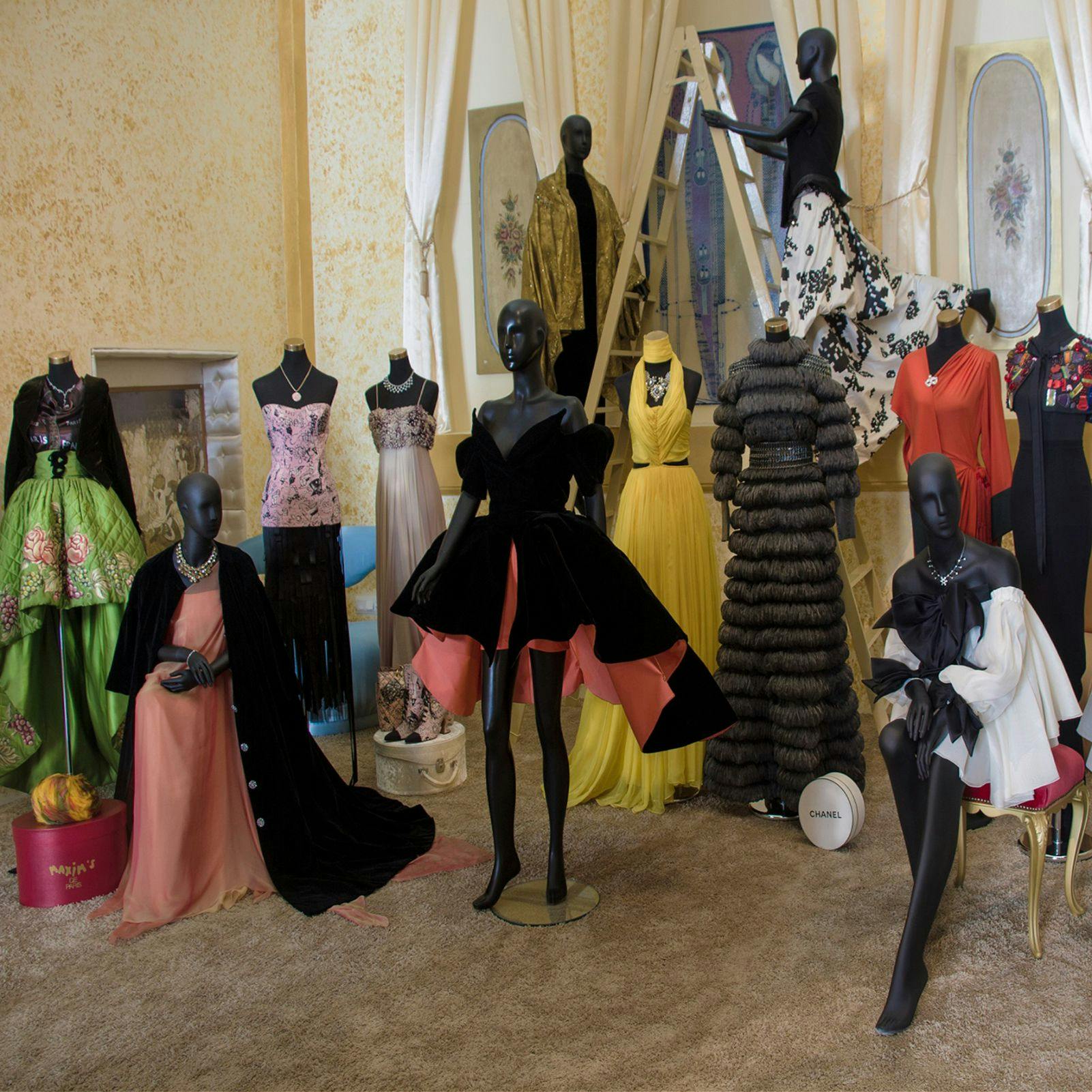 Et rum med flere mannequiner klædt i en række udførlige kjoler, herunder kjoler og formelt slid, på et gulvtæppe.