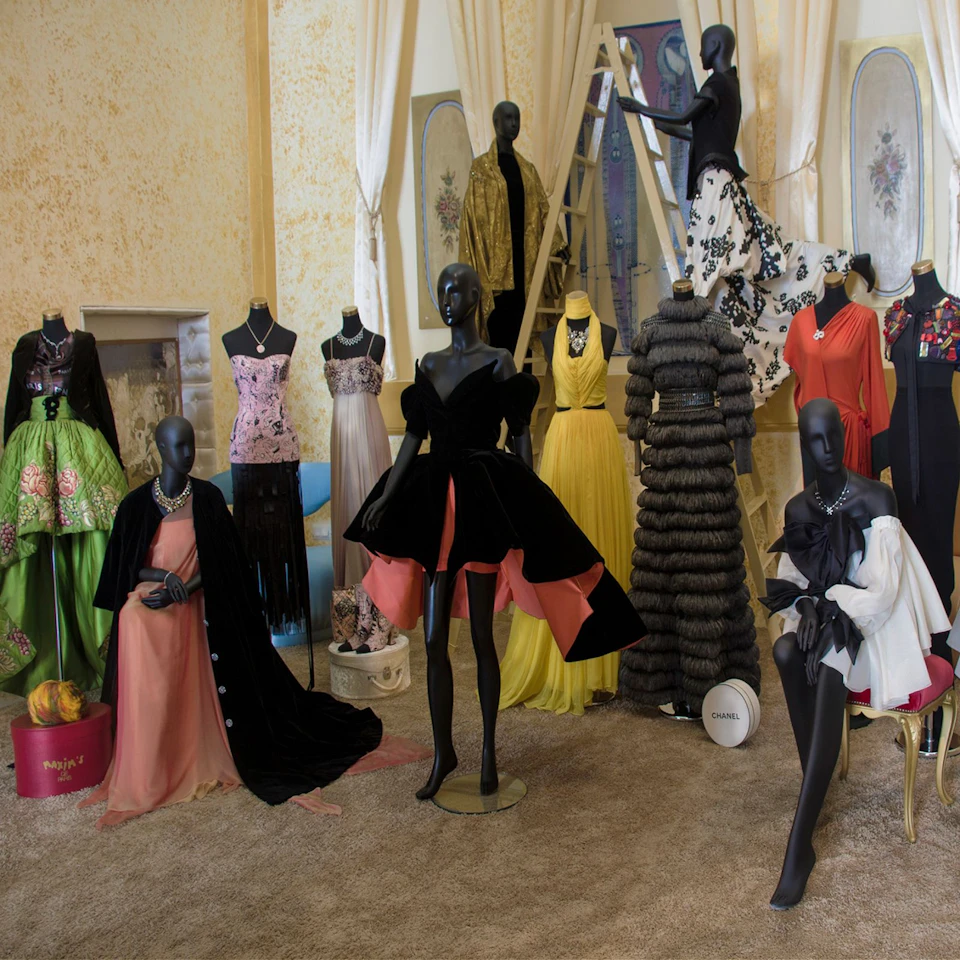 A room with multiple mannequins dressed in a variety of elaborate dresses, including gowns and formal wear, on a carpeted floor.