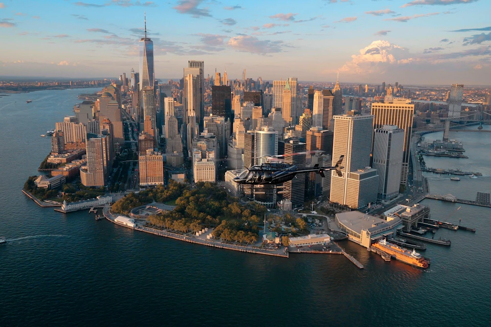 Aerial view of a helicopter flying over the waterfront near skyscrapers in downtown Manhattan at sunset.
