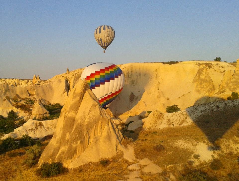 日落时分，两个带有彩虹条纹的热气球飞过多岩石、干旱的景观。