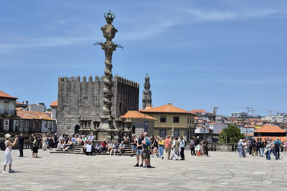 A plaza with a decorative column, people sitting and walking, and historic buildings under a clear blue sky.