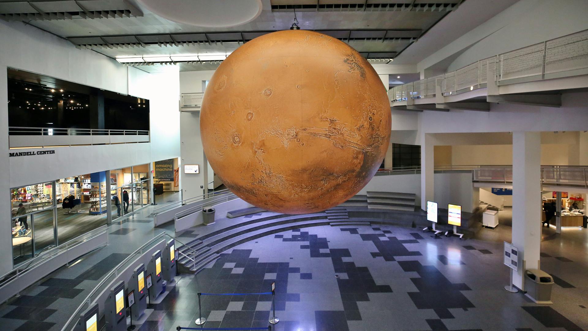 階層状のシーティングエリアとインタラクティブなディスプレイを備えたモダンな屋内スペースの上に、火星の大型モデルが吊り下げられています。