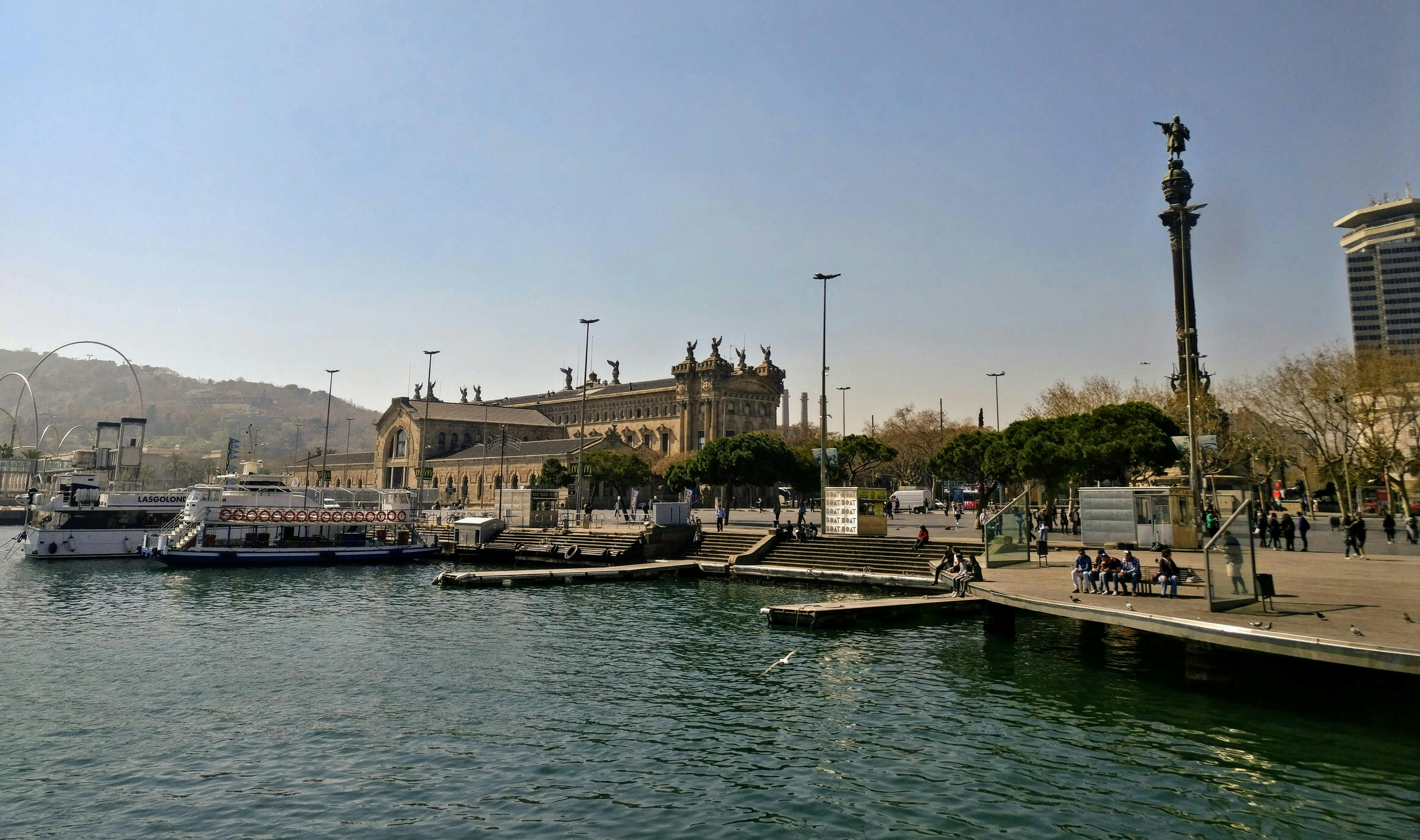 Harbor scene with a historic building, some boats docked, steps leading up from the water, and people walking or sitting nearby.