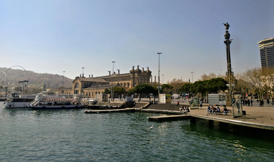 Harbor scene with a historic building, some boats docked, steps leading up from the water, and people walking or sitting nearby.