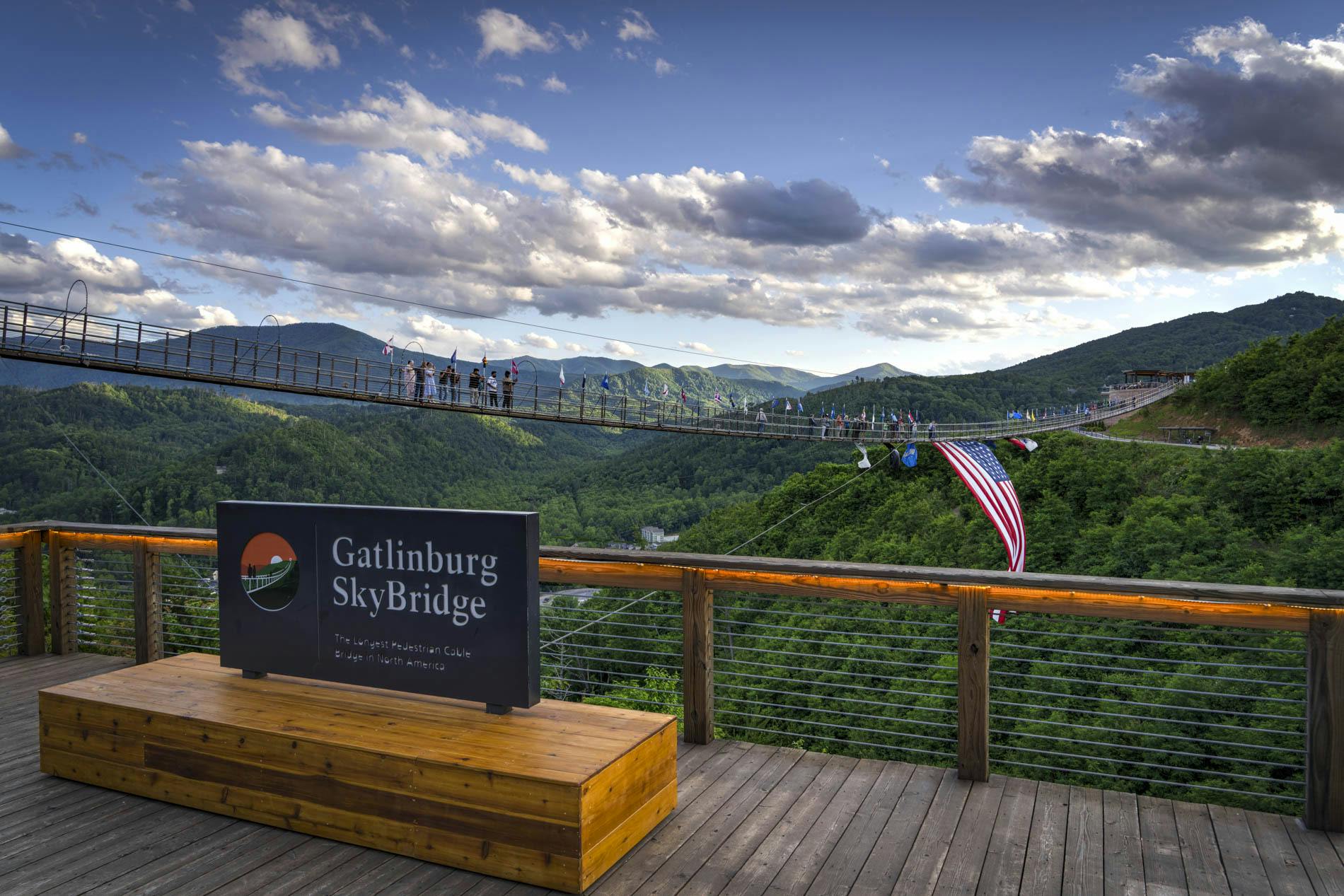 Lidé chodí po Gatlinburg SkyBridge, dlouhém visutém mostě v horách. Na ceduli je nápis „Gatlinburg SkyBridge“.