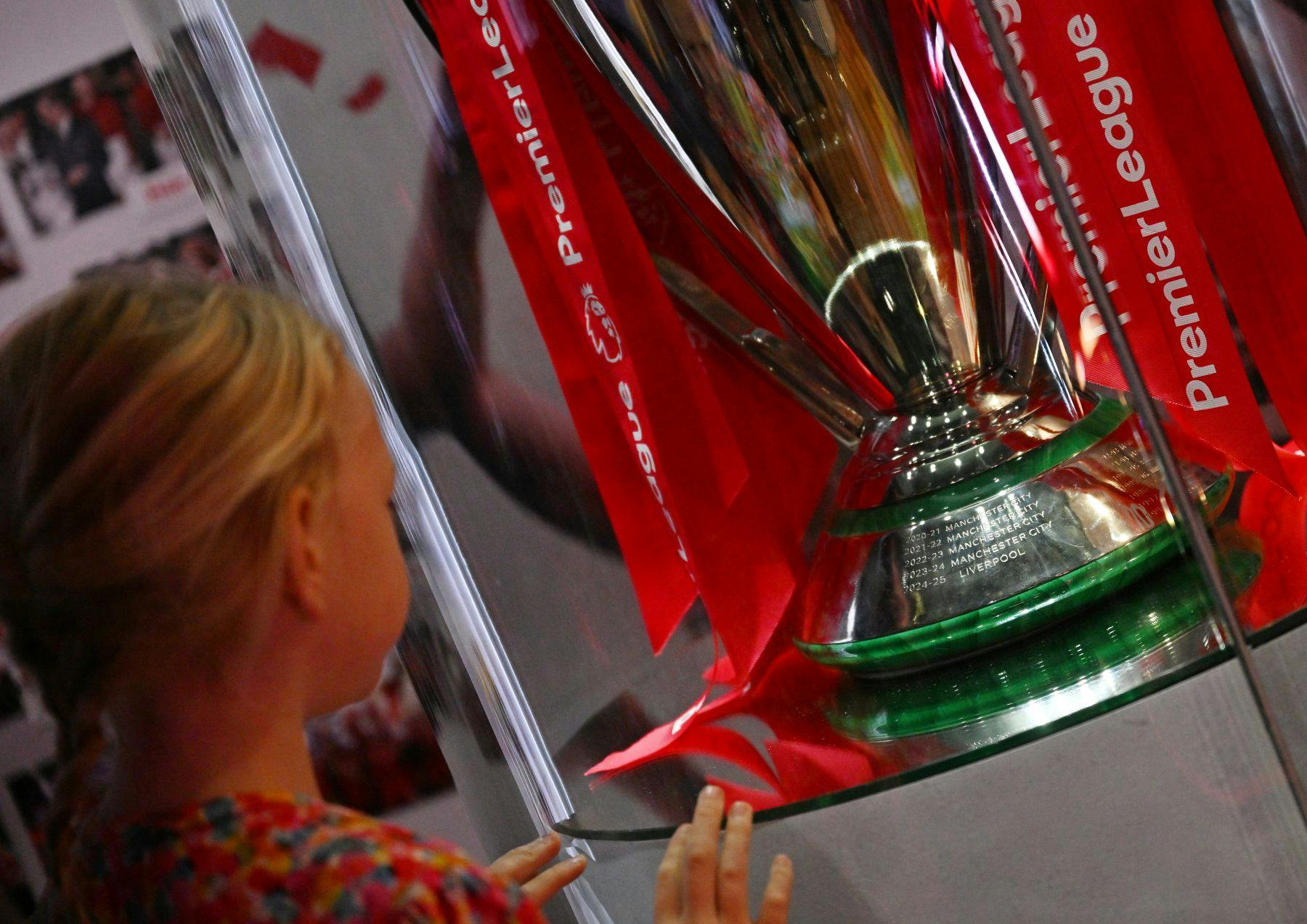 유리 디스플레이 케이스를 통해 빨간색 리본이 달린 프리미어 리그 트로피를 바라보는 아이.