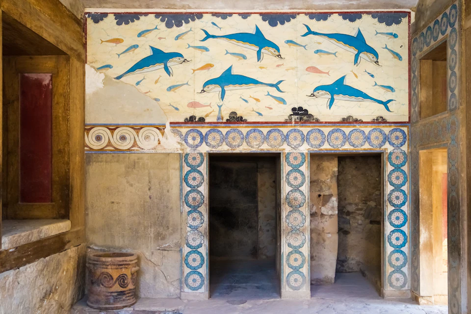 Ancient wall painting of dolphins and fish above three stone doorways with ornate columns and patterns. A pottery jar sits to the side.