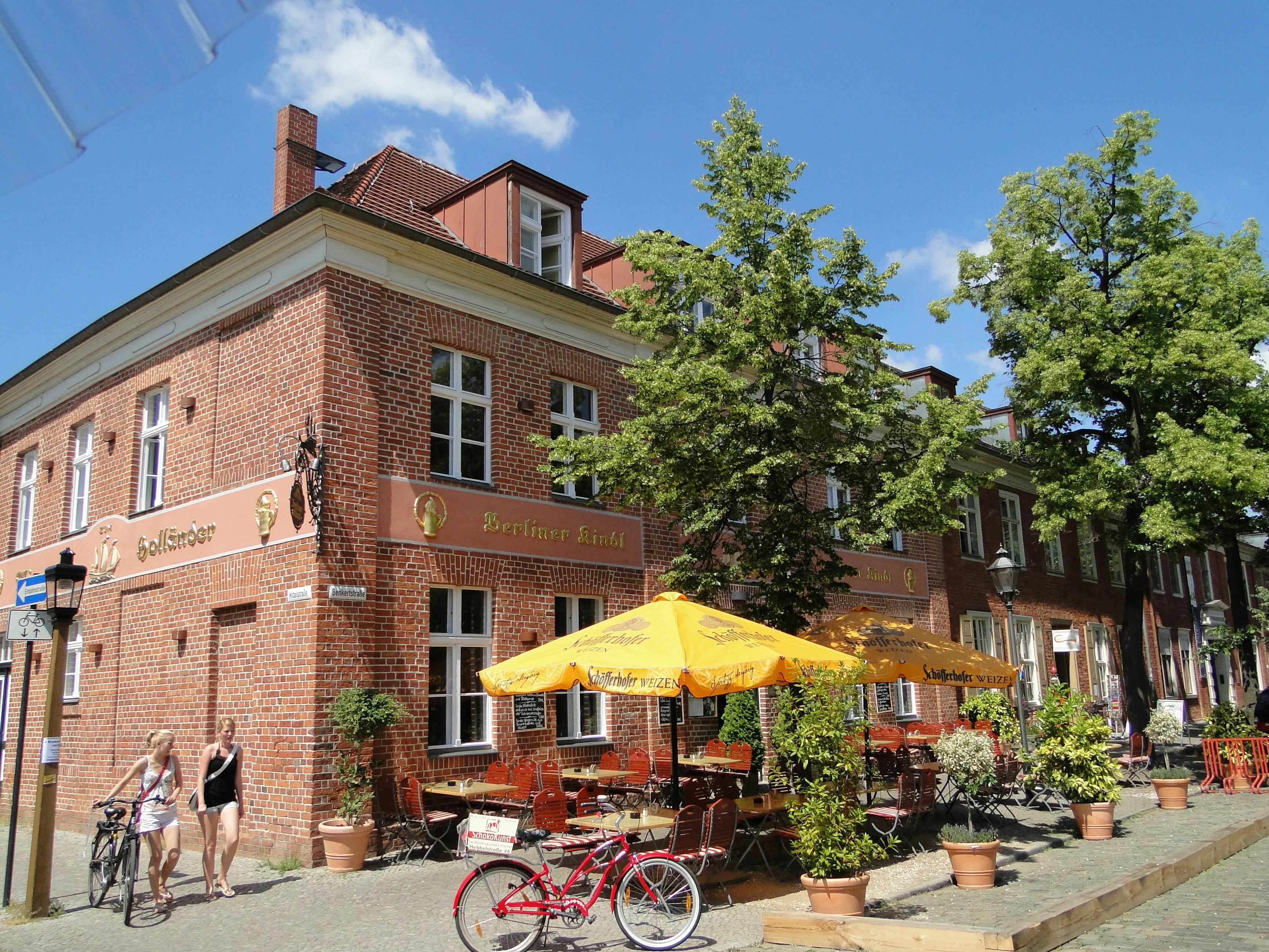 砖砌建筑，黄色雨伞下有户外座椅，绿树成荫，前面停着一辆红色自行车，人们走过。