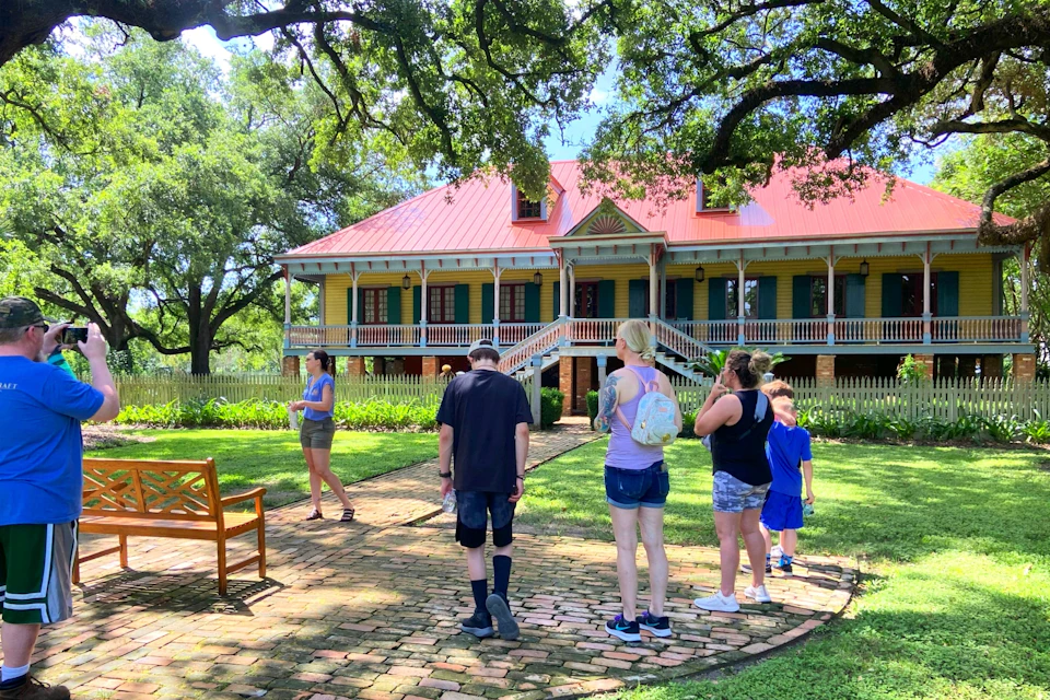 A group of people stand near a path in front of a large, colorful house with a red roof and wraparound porch, surrounded by trees.