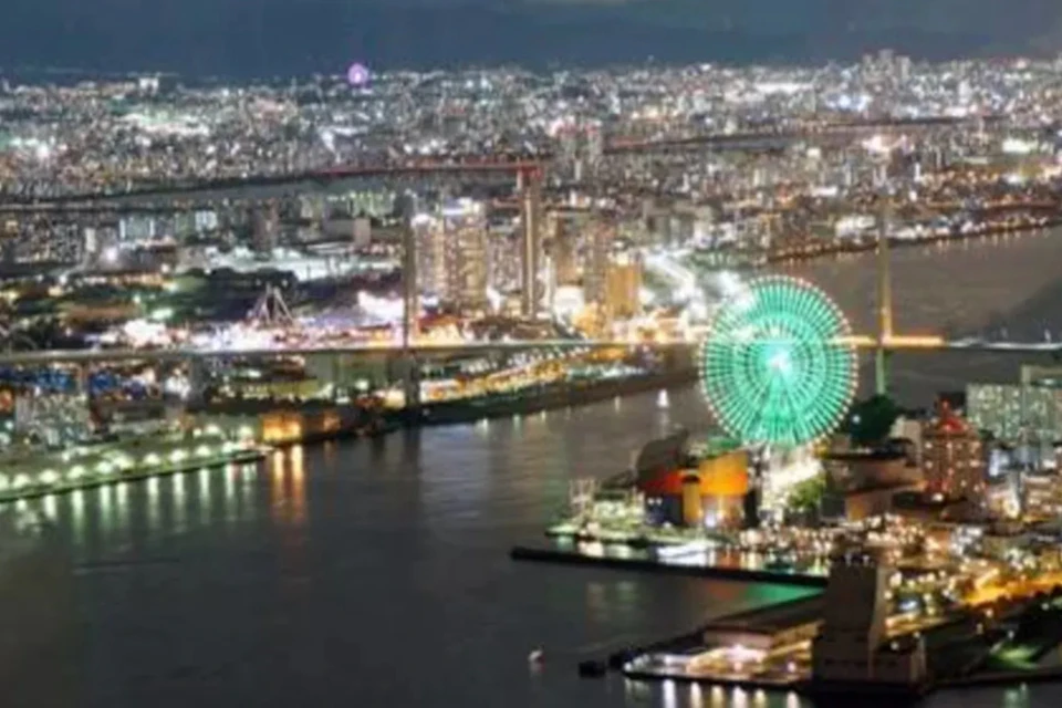 Cityscape at night with a river, illuminated Ferris wheel, and a bridge in the background among brightly lit buildings.