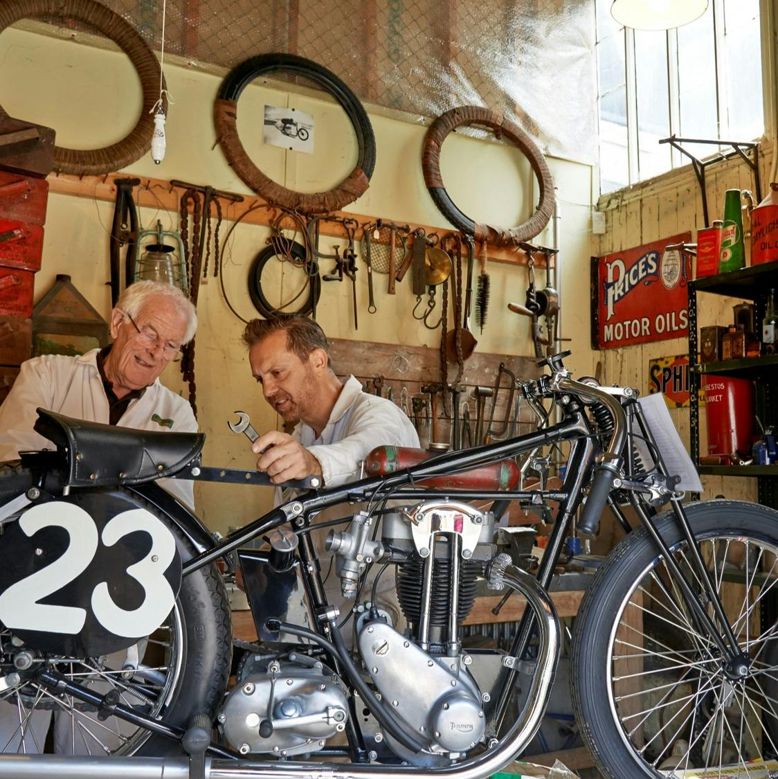 Dos homes treballant en una motocicleta vintage dins d'un taller, amb eines, pneumàtics i cartells d'oli de motor penjats a la paret.