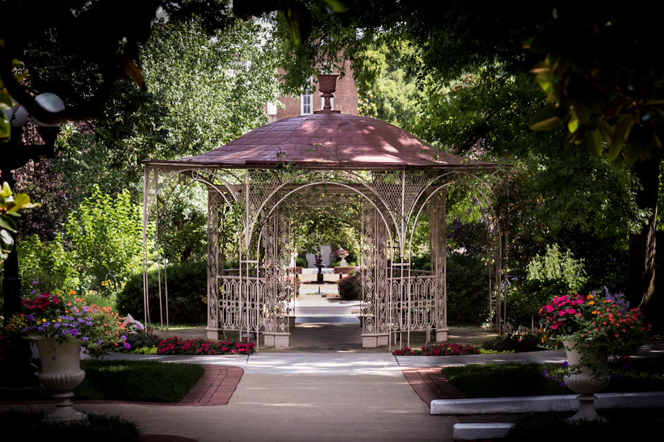 A decorative gazebo with intricate metalwork and a red roof is surrounded by lush greenery and colorful flowers.