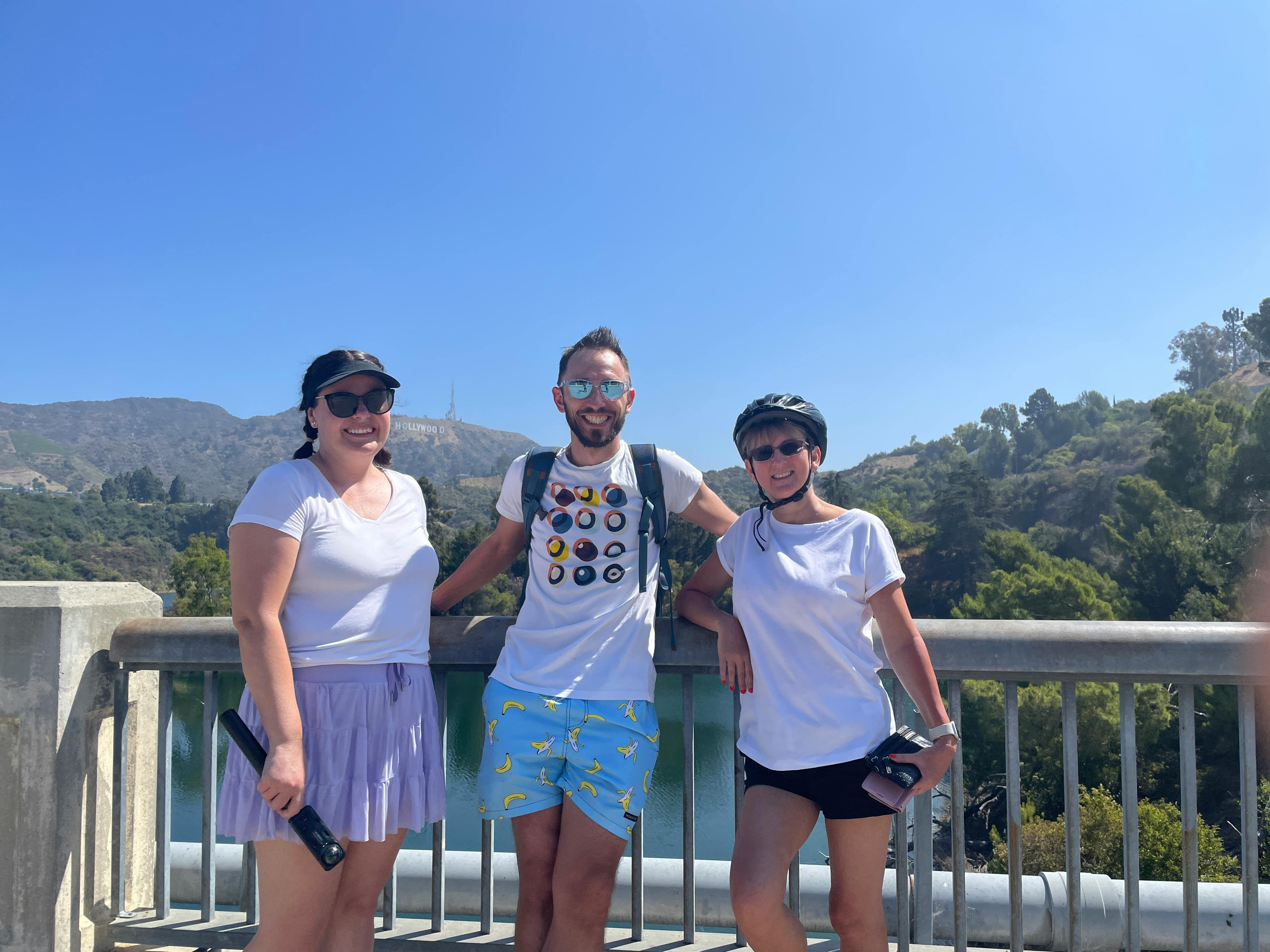 Three people in sporty attire standing on a bridge with a lush, green landscape and clear blue sky in the background.