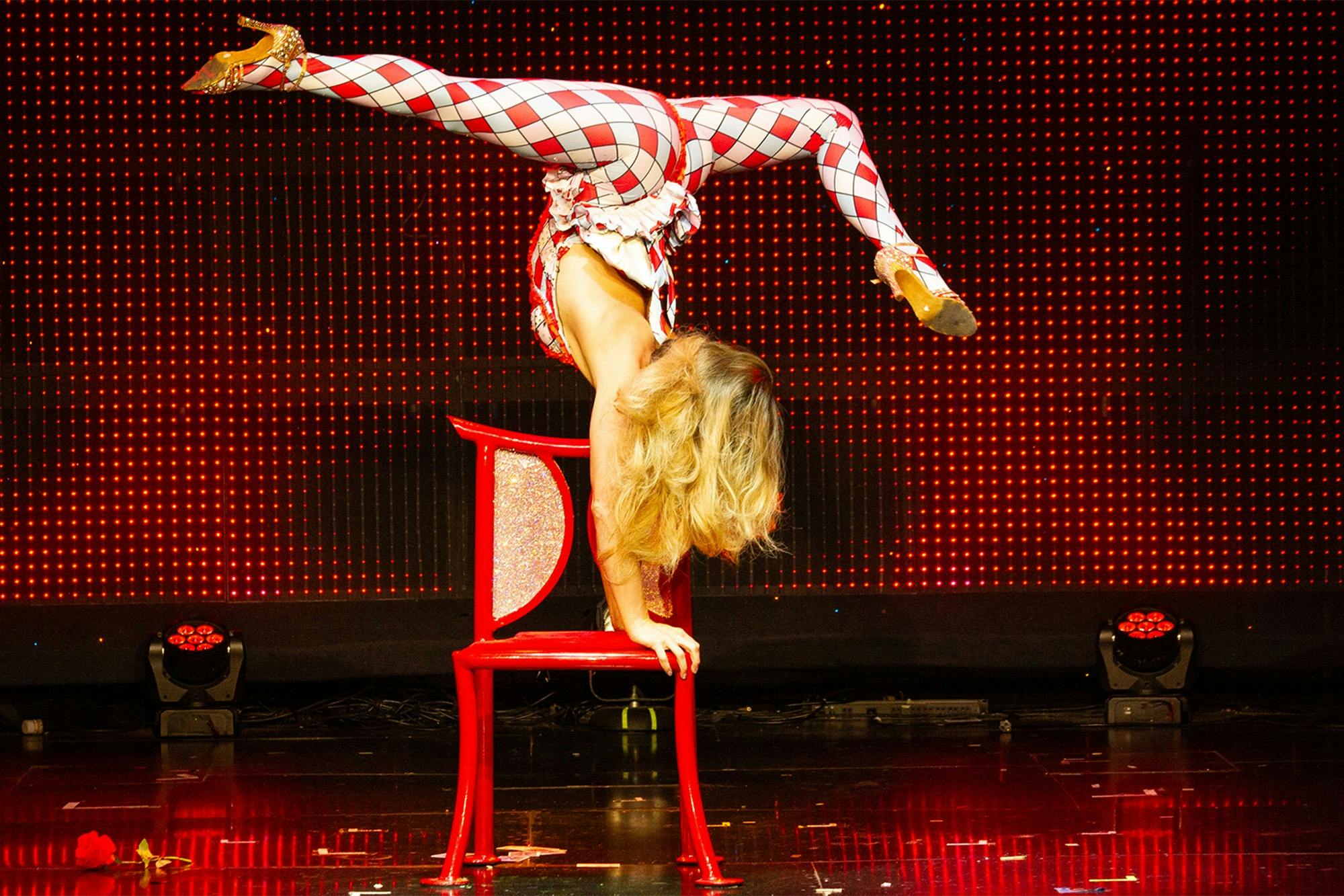 A performer in a red and white harlequin outfit performs a handstand on a red chair against a lit background.
