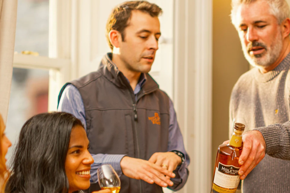 A group of people sit and stand around a wooden table, tasting wine or spirits while talking in a cozy indoor setting.