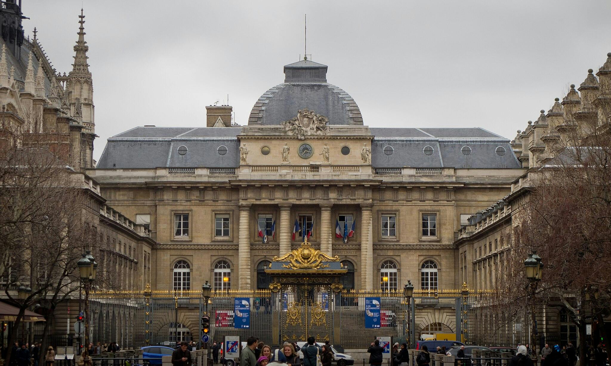 Bâtiment historique à l'architecture ornée, porte dorée et drapeaux multiples. Ciel couvert ; piétons visibles au premier plan.