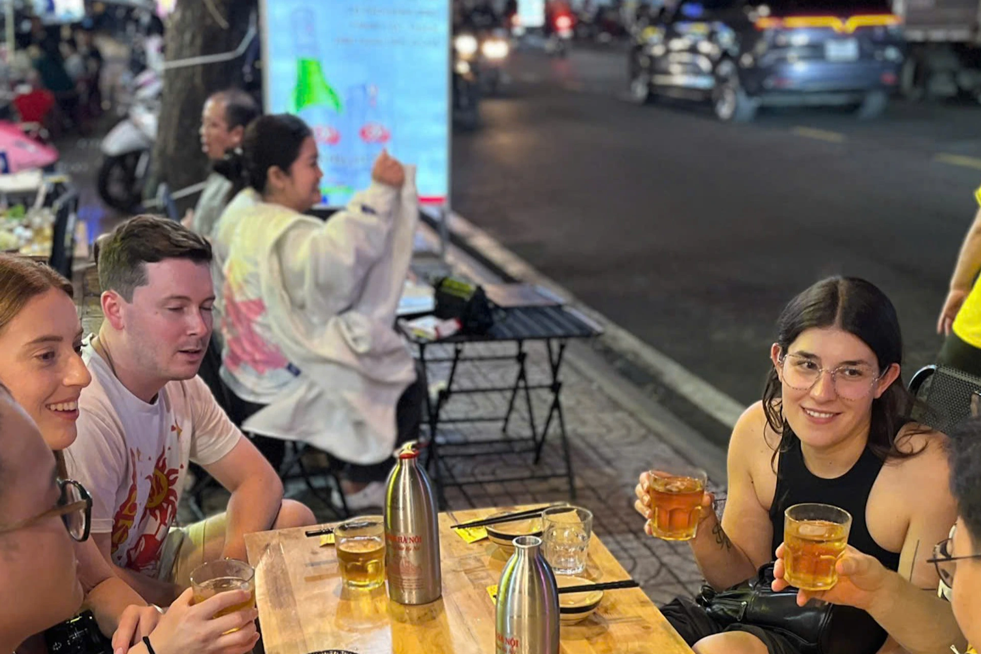 A group of people sit around an outdoor wooden table with food and drinks, engaging in conversation at a busy street-side eatery.