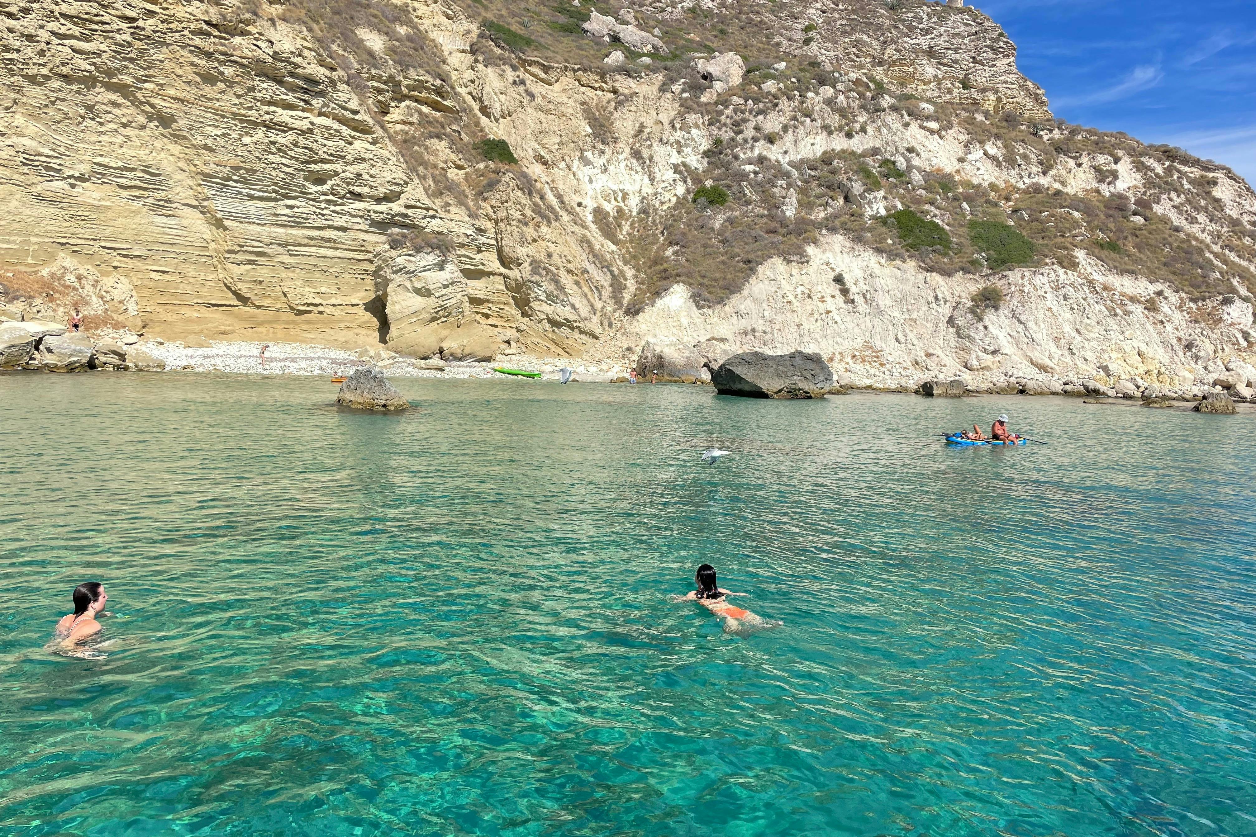 A person swims in crystal-clear turquoise water near a rocky shoreline, with others kayaking in the background.