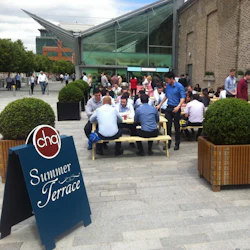 Människor som sitter och står runt picknickbord på en terrass, med en skylt med texten "Summer Terrace" i förgrunden.