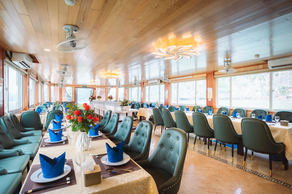 A spacious, elegant dining area with green upholstered chairs, tables set with blue napkins, and floral centerpieces under warm lighting.