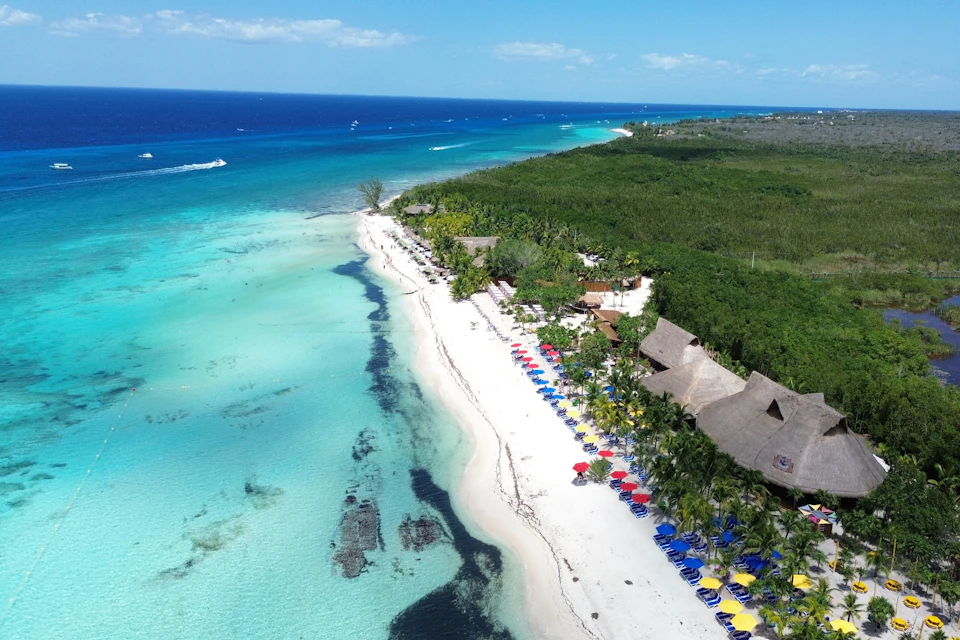 Aerial view of a beach with clear turquoise water, lined with colorful beach umbrellas and chairs, adjacent to green foliage.