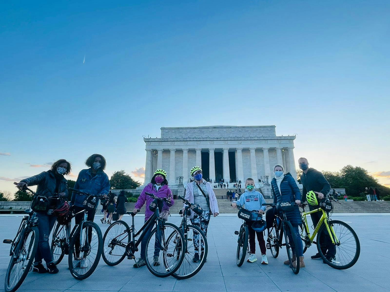 Group of people with bicycles in front of the Lincoln Memorial at dusk, all wearing helmets and masks.