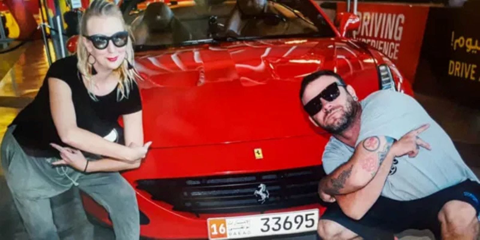 A woman wearing sunglasses and casual clothing poses next to a red sports car with the number 16 on the front plate.