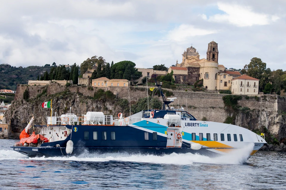 A ferry with the logo 'Liberty Lines' travels on water, with a historic town and church visible on the coast behind it.