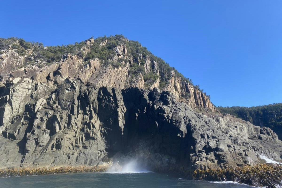 A rocky coastal cliff with vegetation under a clear blue sky, waves splashing against the rocks at the base.