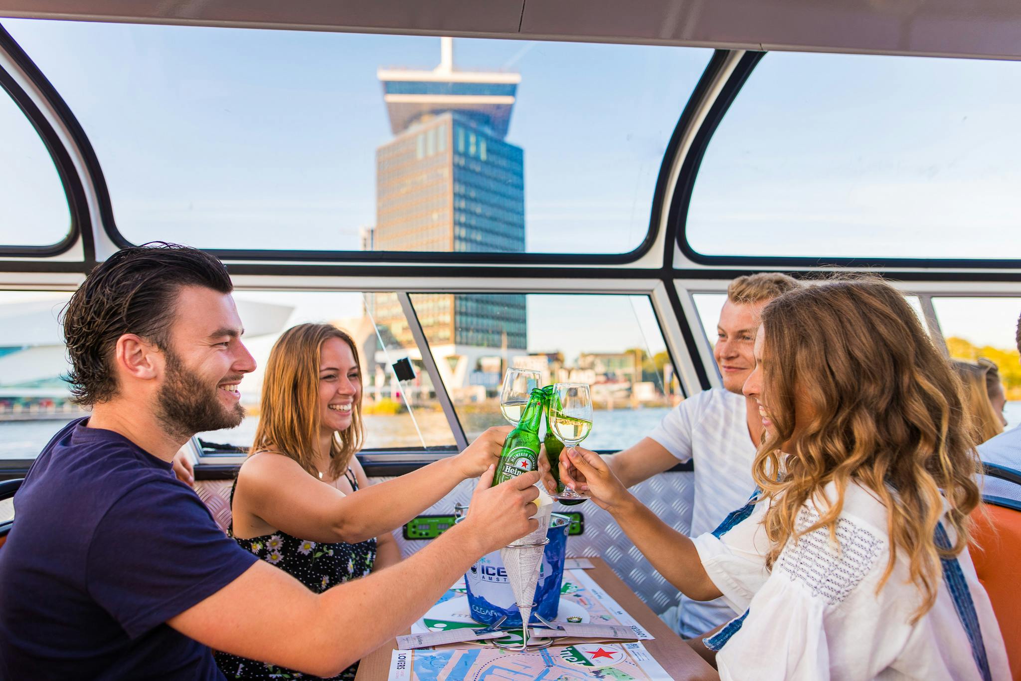 Vier Personen sitzen an einem Tisch in einem Boot und stoßen mit Bierflaschen und Weingläsern an, während durch die Fenster ein Wolkenkratzer zu sehen ist.
