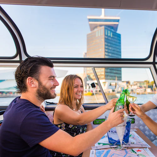 Fyra personer vid ett bord inne i en båt, skålade med ölflaskor och vinglas, med en skyskrapa synlig genom fönstren.