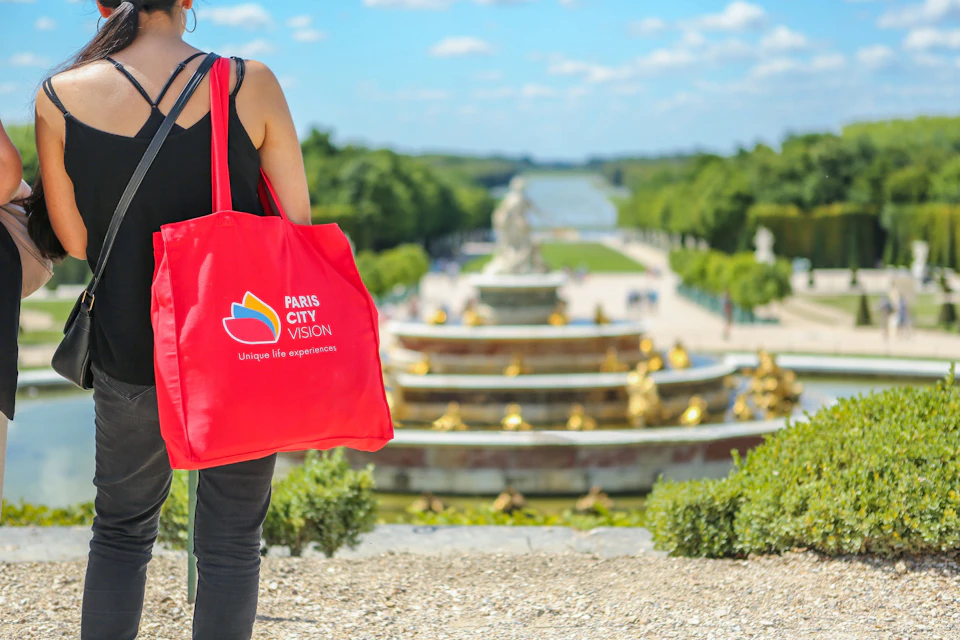 A person with a red tote bag stands near a fountain in a garden with a long water feature in the background.